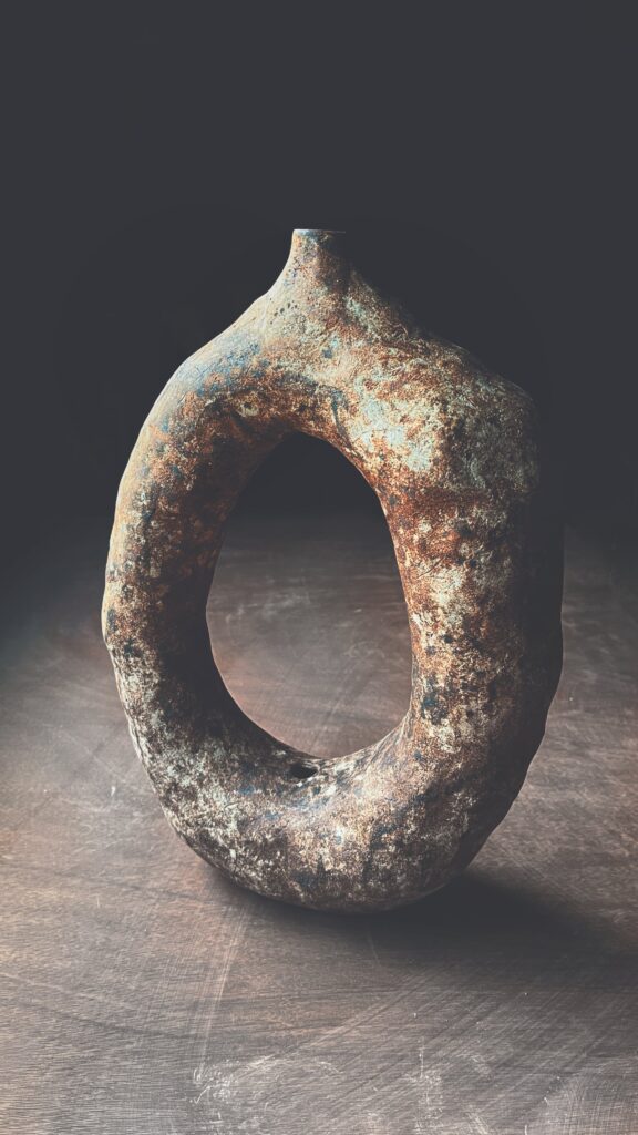 Escultura anular de cerámica primitiva con textura mineral en tonos óxido y verdín