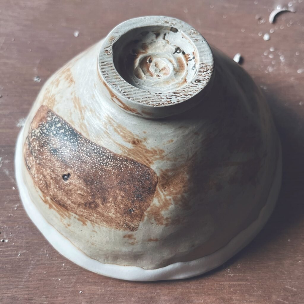 Cuenco de té artesanal visto desde abajo, con esmalte crema y una mancha tostada, y el pie torneado a la vista
